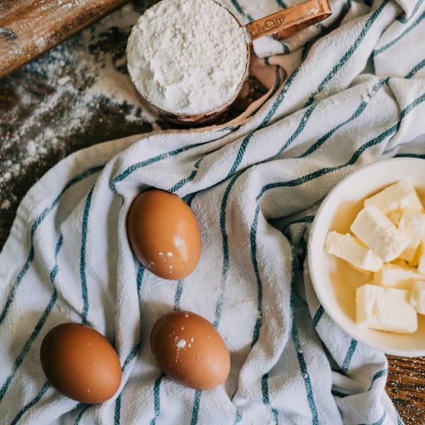 Bowl of cubed butter beside brown eggs and a measuring cup of flour on a striped kitchen towel