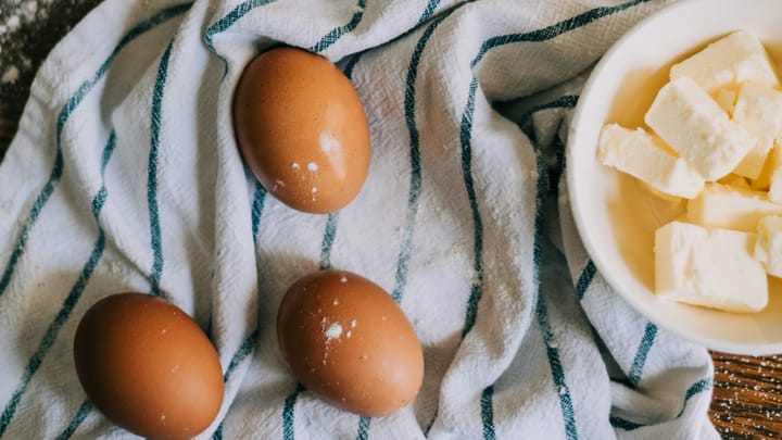 Bowl of cubed butter beside brown eggs and a measuring cup of flour on a striped kitchen towel